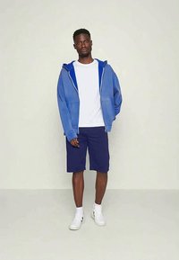 Blå zip-opp hettegenser lagt over en hvit t-skjorte, kombinert med marineblå shorts og hvite sneakers. Enkelt stoff, avslappet passform, og ingen synlige mønstre.