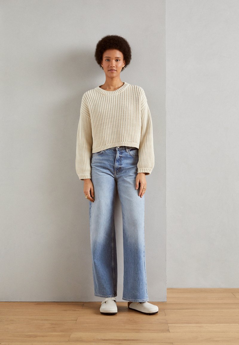 Jersey de punto grueso color beige, pantalones vaqueros de pierna ancha color azul claro y zapatillas blancas sin cordones. Presentado contra una pared gris y un suelo de madera.