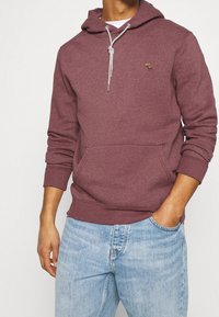 Uomo che indossa una felpa con cappuccio color granata con tasca a marsupio e coulisse, abbinata a jeans denim azzurri, con una mano in tasca.