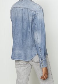 Jasnoniebieska koszula dżinsowa z długimi rękawami, zaokrąglonym dołem i guziczkami na mankietach. Szare spodnie o fakturze z jasnym przetarciem.