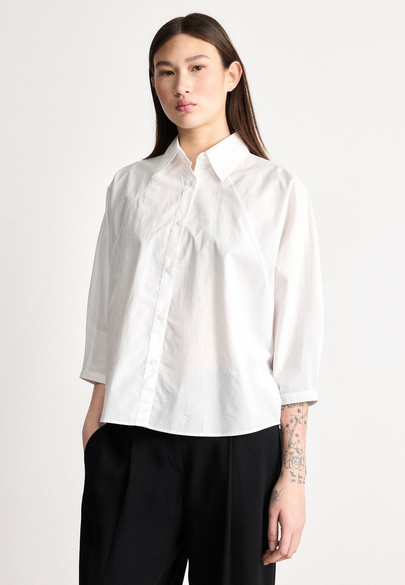 Λευκό πουκάμισο με κουμπιά, με άνοιγμα για γιακά και μανίκια τριών τετάρτων, με άνετη εφαρμογή και μαλακή υφή βαμβακιού, σε συνδυασμό με μαύρο παντελόνι.