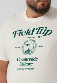 Cremfarbendes T-Shirt aus Baumwolle mit grünem Text und kreisförmigem Grafikdesign, das Vögel und Laub darstellt. Runder Halsausschnitt und kurze Ärmel.