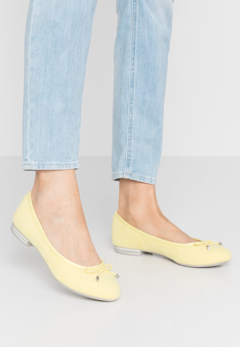 Pieds portant un jean bleu clair et des ballerines jaune pâle avec de petits nœuds sur un sol gris clair et un fond blanc.