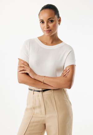 Weißes Strickoberteil mit kurzen Ärmeln und rundem Halsausschnitt, kombiniert mit hoch taillierter beige Hose und einem schwarzen Gürtel. Schlichtes Design, glatte Textur.