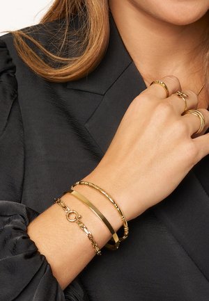 Goudkleurige armbanden met verschillende ontwerpen: een gladde band, een kralenstijl en een ketting met een sluiting, gedragen om een pols.