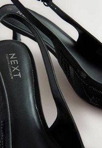 Mustat slingback-korkokengät, joissa on sileä nahkapinta, hienovarainen kohokuviointi ja pehmeä pohjallinen. Hopeinen logo jalkapohjassa.
