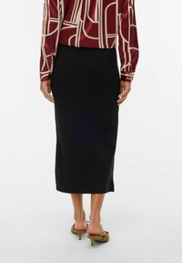 Falda midi negra con una silueta recta, hecha de una tela suave. Combinada con sandalias de tacón marrones. Textura sutil y diseño limpio.