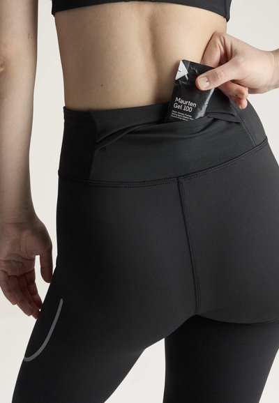 Person iført sorte atletiske leggings placerer Maurten Gel 100 energigel i lommen på bageste linning.