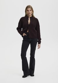 Burgundy, oversized cardigan in a soft, textured wool blend, featuring a stand collar and black buttons. Paired with black flared jeans.