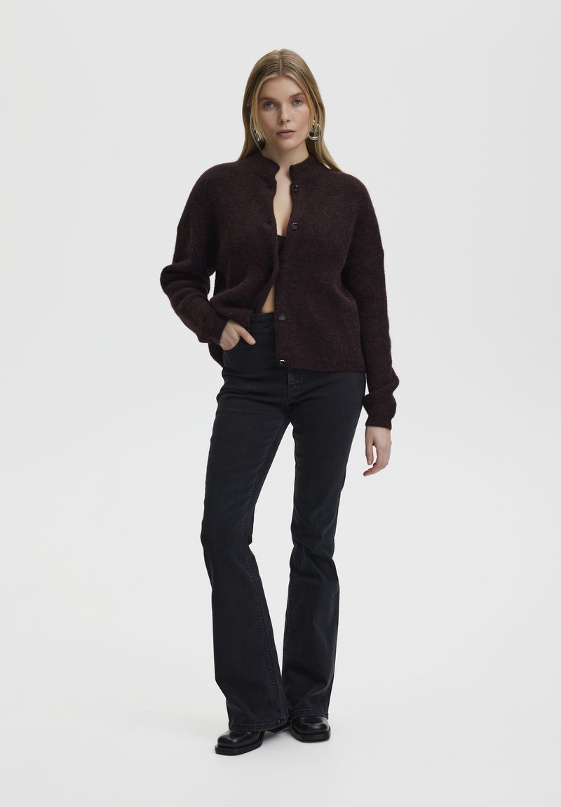 Burgundy, oversized cardigan in a soft, textured wool blend, featuring a stand collar and black buttons. Paired with black flared jeans.