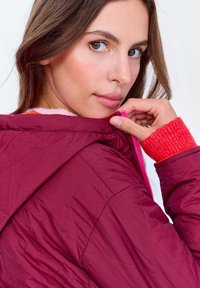 Młoda kobieta trzymająca kołnierz swojego pikowanego burgundowego kurtki, z widocznym różowym zamkiem błyskawicznym i czerwonym swetrem z dzianiny.
