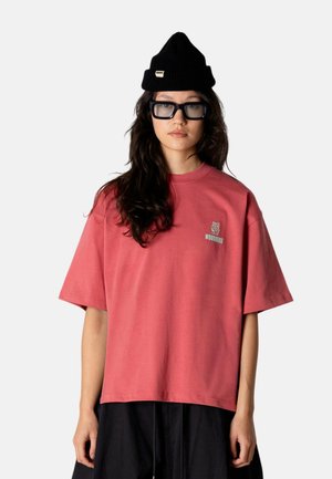Ung person med langt mørkt hår iført en stor lyserød Woodbird T-shirt, sort hue, store sorte briller og sorte bukser, stående.