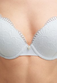 Lichtgrijze kanten bh met een bloemenpatroon, gepadded cups, geschulpte randen en een decoratief detail in goudkleurig metaal in het midden op de rug.
