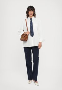 Chemise blanche à manches longues, cravate bleu marine à motifs, pantalon bleu marine et chaussures beiges à enfiler. Sac en bandoulière marron avec des accents en métal doré.