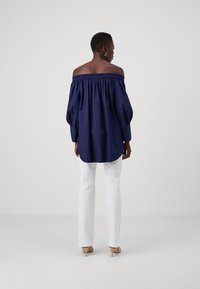 Top blu navy a spalle scoperte con scollatura arricciata, maniche lunghe a sbuffo e orlo curvo, abbinato a pantaloni bianchi slim-fit e tacchi nudi.
