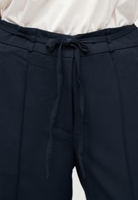 Κοντινό πλάνο μπλε παντελονιού ναυτικού με δεμένη υφασμάτινη ζώνη και κάθετες ραφές, φορεμένο με λευκό μπλουζάκι που έχει μαζευτεί μέσα.