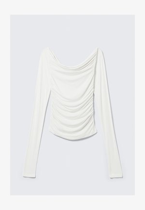 Haut blanc à manches longues, ajusté, avec un effet froncé sur le devant et un large col drapé sur un fond gris clair.