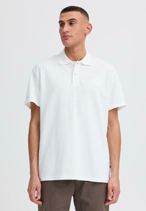 Jeune homme portant un polo blanc à manches courtes uni et un pantalon marron, debout devant un fond clair uni, regardant sur le côté.