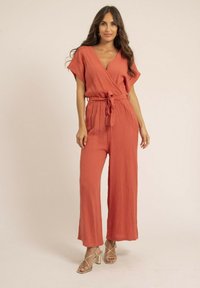 Combinaison en terracotta à manches courtes, ceinture à nouer, jambes larges et tissu texturé. Associée à des talons dorés à lanières.