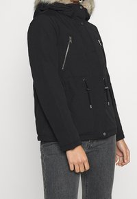 Svart vinterjacka med huva och pälsdetalj, med dragkedjefickor på bröstet, dragsko i midjan, bärs med gråa jeans på en stående person.