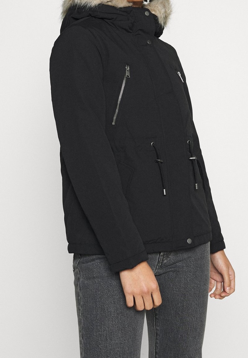 Svart vinterjacka med huva och pälsdetalj, med dragkedjefickor på bröstet, dragsko i midjan, bärs med gråa jeans på en stående person.