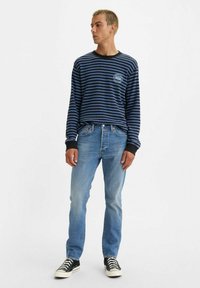 Camisa de manga larga a rayas azules y negras con cuello negro, logo en el pecho, combinada con vaqueros azul claro y zapatillas negras.