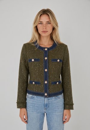 Vrouw met lang blond haar draagt een olijfgroene getextureerde jas met blauwe bies en gouden knopen, gecombineerd met lichtblauwe spijkerbroek, staat stil.