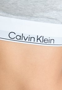 Cintura de tela gris con ribete blanco, con el logo "Calvin Klein" en negro. Textura suave y diseño aerodinámico.