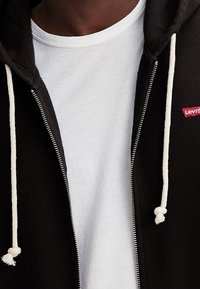 Zwart zip-up hoodie met crèmekleurige trekkoord over een wit t-shirt. Bevat een klein rood Levi's-logo aan de linkerkant. Zachte stofstructuur.