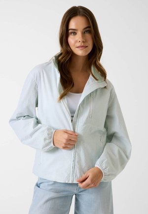 Junge Frau mit langen braunen Haaren, die eine hellblaue, strukturierte Jacke mit Reißverschluss über einem weißen Oberteil und hellblauen Jeans trägt, und den Reißverschluss der Jacke anpasst.