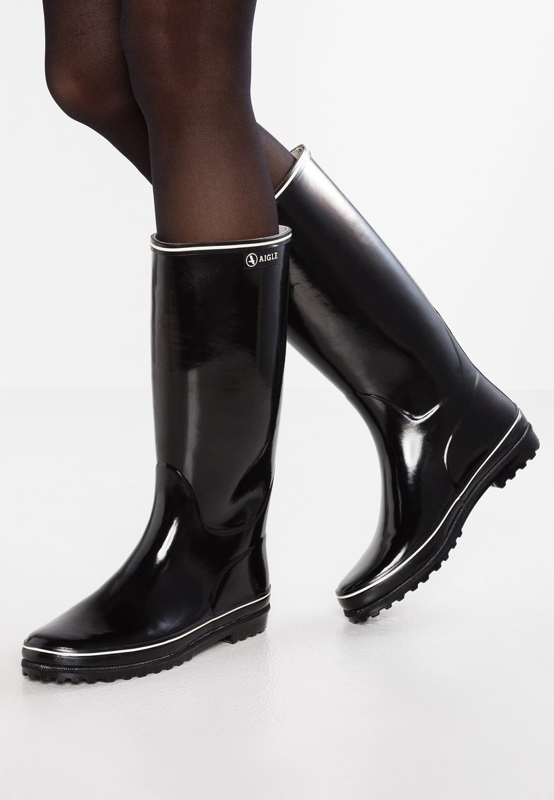 Bottes noires en caoutchouc montantes jusqu'au genou, avec une finition brillante, un bord argenté en haut, et une semelle noire texturée pour l'adhérence. Logo Aigle visible.
