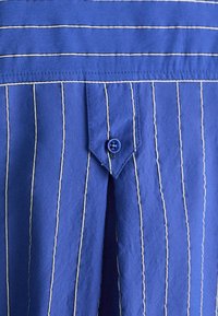 Camicia di stoffa a righe blu con strisce bianche alternate e strette, taschino a punta e dettaglio blu sui bottoni. Materiale in cotone morbido e testurizzato.