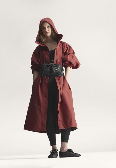 Donna che indossa un lungo cappotto con cappuccio color ruggine, leggings neri, sneakers nere e una cintura nera con logo bianco.