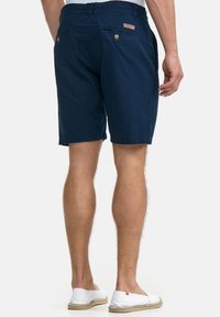 Marineblaue Baumwollshorts mit zwei Gesäßtaschen und goldenen Knöpfen, gerader Schnitt und glatter Struktur. Dazu weiße Slip-on-Schuhe getragen.