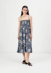 Blå blommig midiklänning med tunna band och smockad byst. Har vita blommönster och en utsvängd kjol. Bärs med svarta sandaletter med öppen tå.