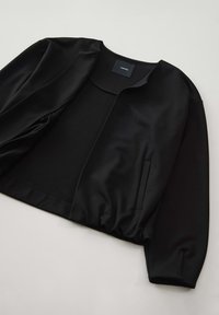 Veste noire minimaliste en tissu lisse et extensible, à col rond et dotée de deux poches latérales. Le design présente une coupe décontractée avec des manches froncées.