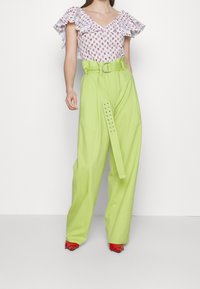Blusa bianca a fiori con maniche a volant abbinata a pantaloni verde lime a vita alta con dettaglio di fibbia e cintura, e scarpe rosse.