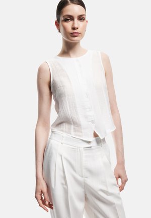 Femme portant un haut blanc sans manches boutonné et un pantalon blanc plissé taille haute, debout devant un fond clair uni.