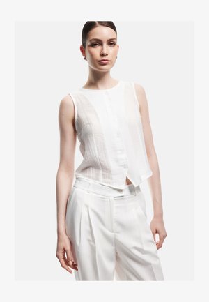 Femme portant un haut blanc sans manches boutonné et un pantalon blanc plissé taille haute, debout devant un fond clair uni.