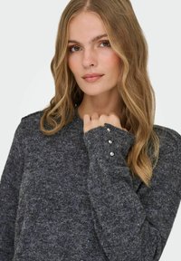 Sivi, teksturirani pulover s dugim rukavima. Manžetna ima dekorativne srebrne gumbe, a izrez je okrugli. Mekana tkanina s udobnim izgledom.