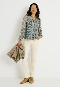 Blouse à manches longues bleu à fleurs avec un devant boutonné, associée à un pantalon évasé crème et des bottines à talons blocks en tan. Tenant un pull beige.