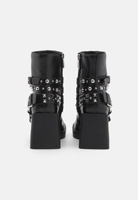 Des bottes à talons en cuir noir présentent un talon carré épais, un zip et plusieurs sangles ornées de rivets argentés et d'accents en forme de pyramide.