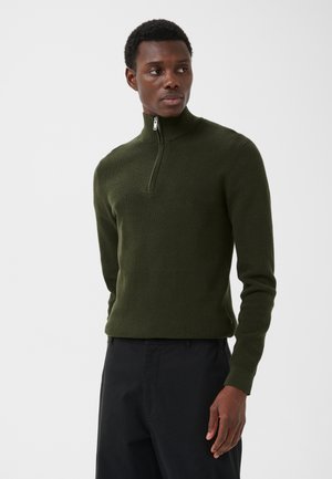 Mann iført en mørkegrønn genser med halv glidelås og svarte bukser, stående mot en ensfarget hvit bakgrunn, ser mot venstre.