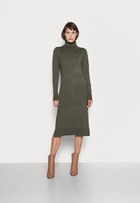 Robe col roulé kaki en tissu tricot doux, design ajusté, manches longues, fentes sur les côtés, associée à des bottines beiges.