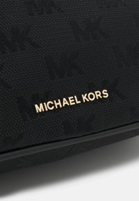 Tessuto nero testurizzato con motivo ripetuto in rilievo "MK" e lettere dorate "MICHAEL KORS" al centro su una superficie piana.