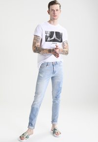White short-sleeve T-shirt with a black graphic print, paired with light blue distressed denim jeans featuring patchwork details and casual sandals.