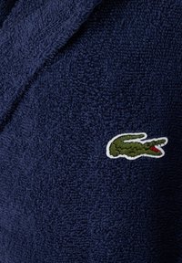 Accappatoio in spugna blu navy con un logo di coccodrillo verde e dettagli rossi, posizionato su una superficie testurizzata. Tessuto morbido e spesso.