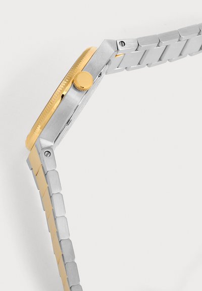 Vue de côté d'une montre-bracelet avec un bracelet en métal argenté et une lunette et couronne en or sur fond blanc.