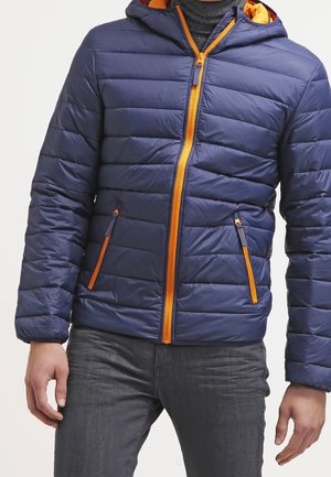 Homme portant une veste matelassée bleue avec une fermeture éclair orange et des détails de poche, associée à un pantalon gris foncé.