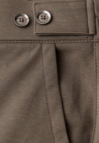 Close-up of brown fabric pants showing two silver buttons labeled "Street One" and stitched pocket details.